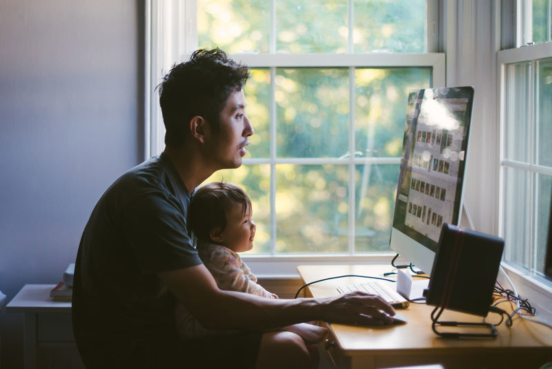 Man working from home at a desk by a window with a baby on his lap